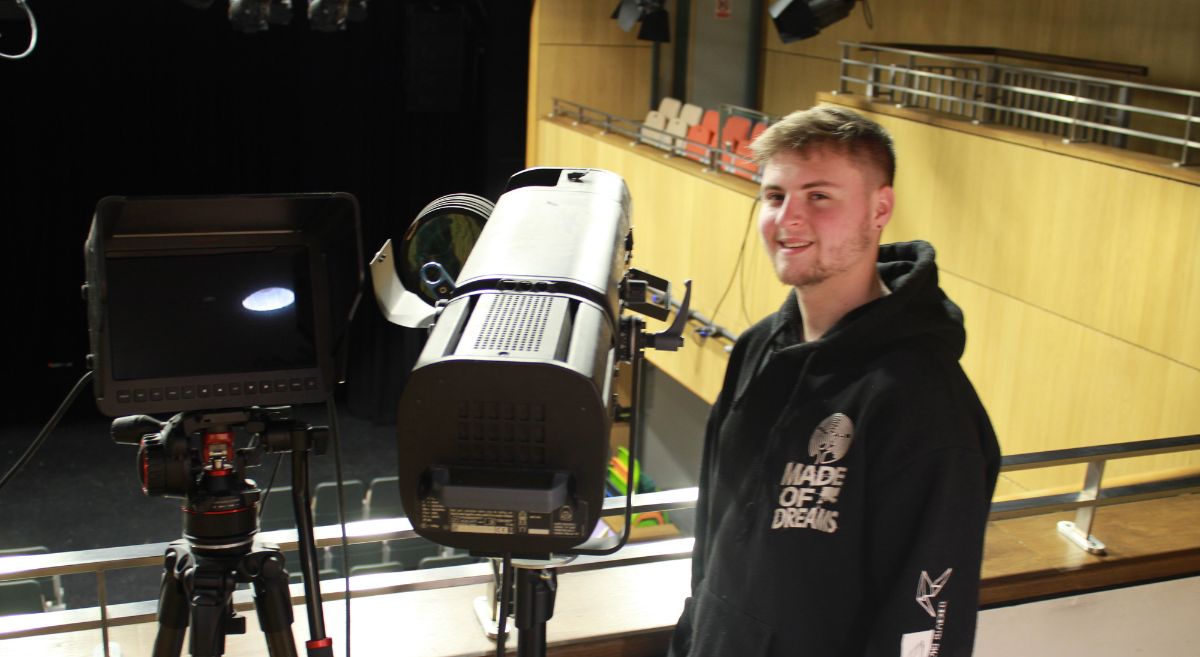 Male student wearing black hoodie standing to left of theatre light and camera monitor with stage in background.